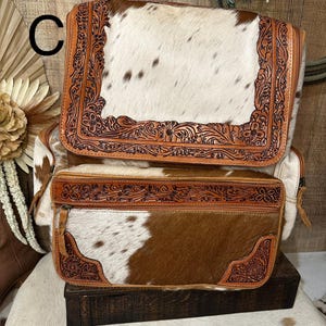 May include: A backpack featuring a white and brown cowhide pattern with tan leather trim. The leather sections are decorated with intricate floral tooling. The bag includes a large top flap and a rectangular front pocket, both secured with zippers.