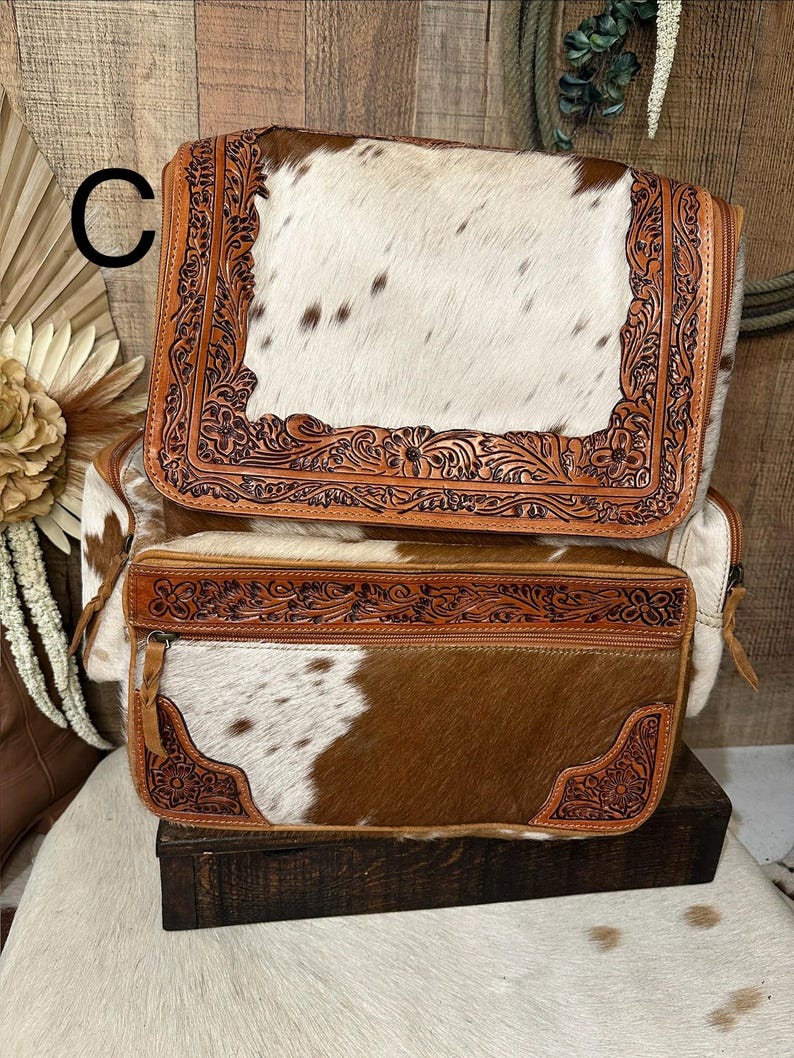 May include: A backpack featuring a white and brown cowhide pattern with tan leather trim. The leather sections are decorated with intricate floral tooling. The bag includes a large top flap and a rectangular front pocket, both secured with zippers.