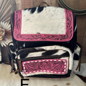 May include: A backpack featuring a black and white cowhide pattern with bright pink leather accents. The top flap and front pocket are decorated with intricate floral tooling and white stitching. The bag includes side pockets and brown zipper pulls.