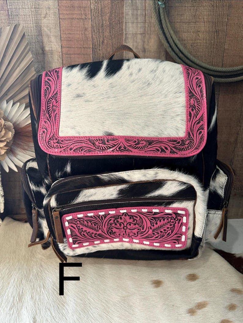 May include: A backpack featuring a black and white cowhide pattern with bright pink leather accents. The top flap and front pocket are decorated with intricate floral tooling and white stitching. The bag includes side pockets and brown zipper pulls.