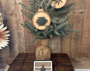 May include: A small Christmas tree decorated with wooden ornaments. The ornaments feature sun and geometric designs. A rectangular ornament with a Christmas tree design is in the foreground. The tree is on a wooden stand.