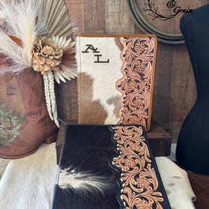 May include: Two rectangular zippered portfolios featuring a combination of hair-on-hide leather and hand-tooled floral leather panels. The top portfolio is tan and white with the initials AL, while the bottom portfolio is black and white.