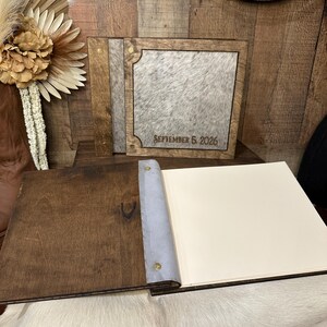 Peut inclure: Un album photo carr&eacute; avec un cadre en bois brun fonc&eacute; et un panneau central en peau de vache grise et blanche. La couverture porte le texte grav&eacute; SEPTEMBER 5, 2026. L'album ouvert pr&eacute;sente une couverture en bois brun, une reliure grise et des pages cr&egrave;me.