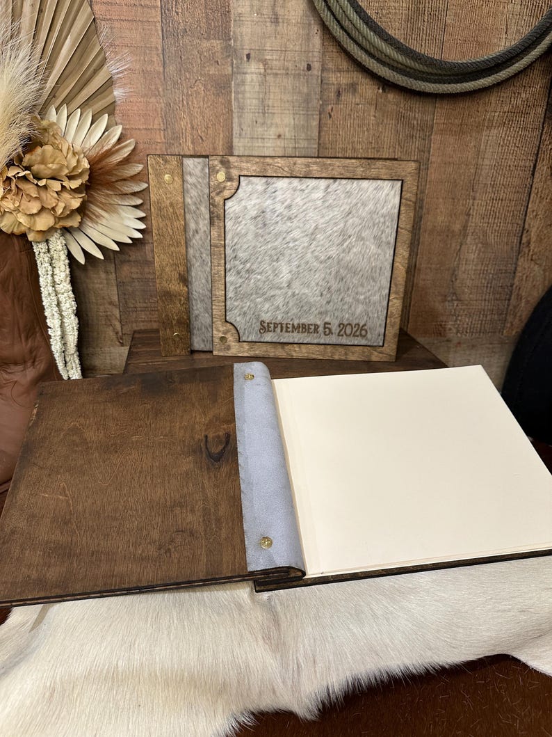 Peut inclure: Un album photo carr&eacute; avec un cadre en bois brun fonc&eacute; et un panneau central en peau de vache grise et blanche. La couverture porte le texte grav&eacute; SEPTEMBER 5, 2026. L'album ouvert pr&eacute;sente une couverture en bois brun, une reliure grise et des pages cr&egrave;me.