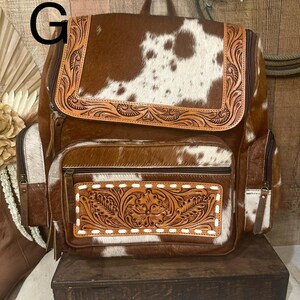 May include: A brown and white cowhide backpack featuring a top flap and a front pocket with intricate floral leather tooling. The bag includes side pockets, brown leather trim, and a top handle, with the letter G visible in the upper left corner.