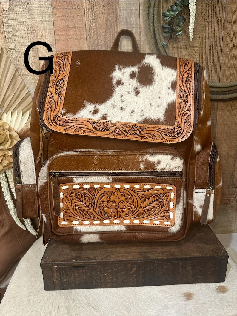 May include: A brown and white cowhide backpack featuring a top flap and a front pocket with intricate floral leather tooling. The bag includes side pockets, brown leather trim, and a top handle, with the letter G visible in the upper left corner.