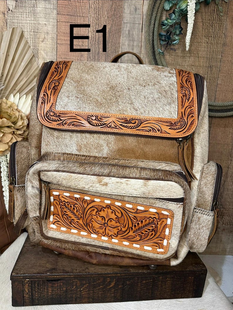 Op de afbeelding: Een rugzak gemaakt van beige en witte koeienhuid met bruine lederen accenten en gedetailleerd bloemmotief. De tas heeft een bovenflap, een voorvak met rits en witte stiksels, en zijvakken, allemaal afgesloten met bruine ritsen.