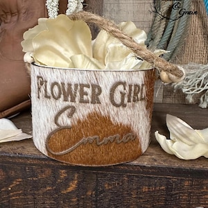 Peut inclure: Un panier cylindrique recouvert de peau de vache brune et blanche avec une anse en corde de jute et des perles en bois. L'avant est gravé avec le texte FLOWER GIRL et le prénom Emma en écriture cursive. Le panier contient des pétales en tissu blanc.