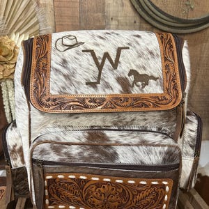 Op de afbeelding: Een rugzak van wit en bruin koeienhuid met een bovenflap met een rand van bewerkt leer, een geborduurde cowboyhoed, de letter W en een rennend paard. Het voorvak bevat een rechthoekig paneel van bewerkt leer met witte stiksels.