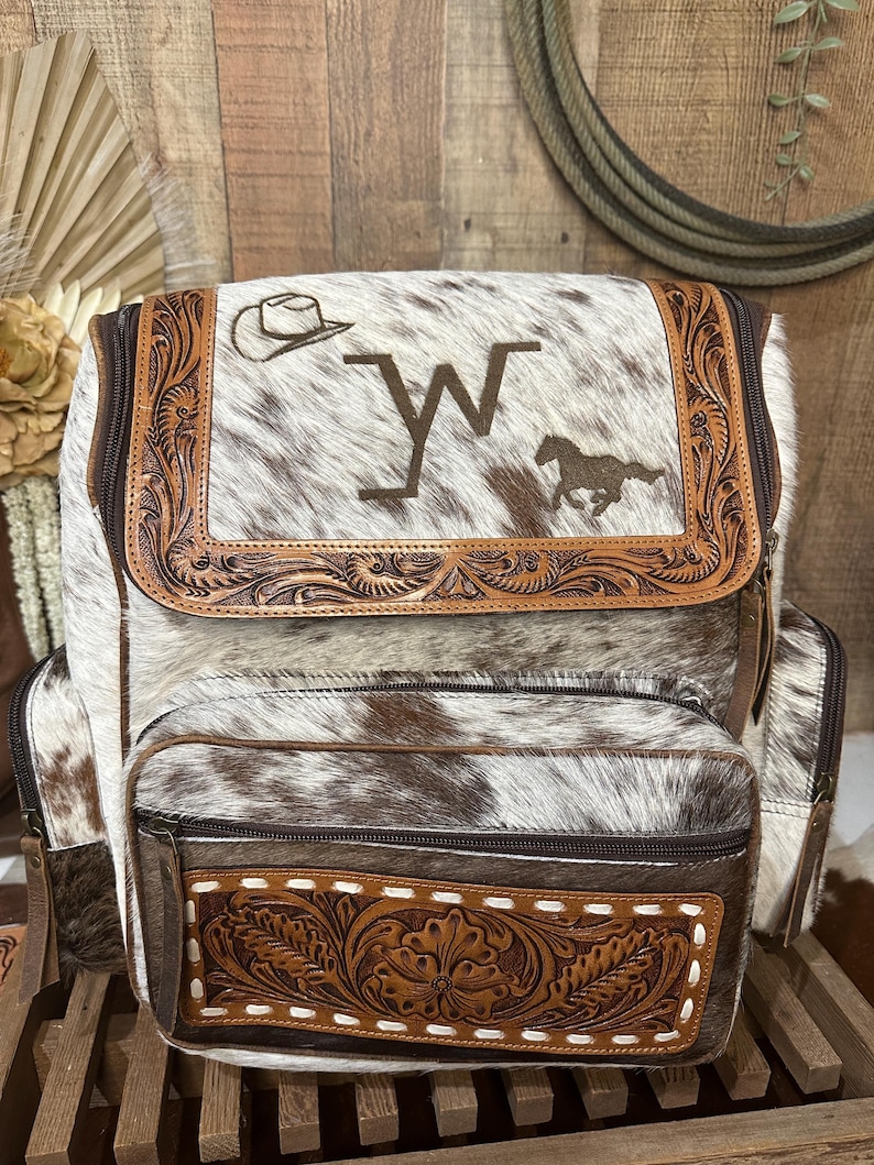 Op de afbeelding: Een rugzak van wit en bruin koeienhuid met een bovenflap met een rand van bewerkt leer, een geborduurde cowboyhoed, de letter W en een rennend paard. Het voorvak bevat een rechthoekig paneel van bewerkt leer met witte stiksels.