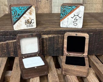 Op de afbeelding: Twee vierkante houten ringdoosjes met deksels in een diagonaal gespleten ontwerp van turquoise reliëfleer en witte vacht. Eén deksel toont de tekst H&C 10-18-25, de andere een symbool. Twee open doosjes tonen witte en zwarte fluwelen inzetstukken.