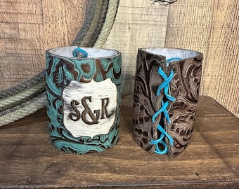 Op de afbeelding: Twee cilindrische lederen hoezen met reliëf bloemmotieven. De linker hoes is turquoise met een wit stukje harig leer met de letters S&R. De rechter hoes is donkerbruin en voorzien van fel turquoise veters in een korsetstijl.