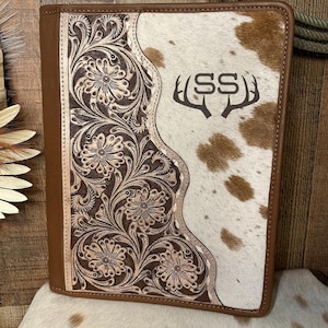 May include: A rectangular leather portfolio featuring a dark brown panel with intricate floral tooling on the left and a white and brown cowhide panel on the right. The cowhide section displays a dark brown logo with the letters SS and antlers.