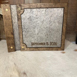 Peut inclure: Un album photo carr&eacute; en bois avec un cadre teint&eacute; brun fonc&eacute; et une incrustation en peau de vache tachet&eacute;e grise et blanche. La couverture pr&eacute;sente le texte grav&eacute; SEPTEMBER 5, 2026, et quatre petits rivets m&eacute;talliques couleur laiton.