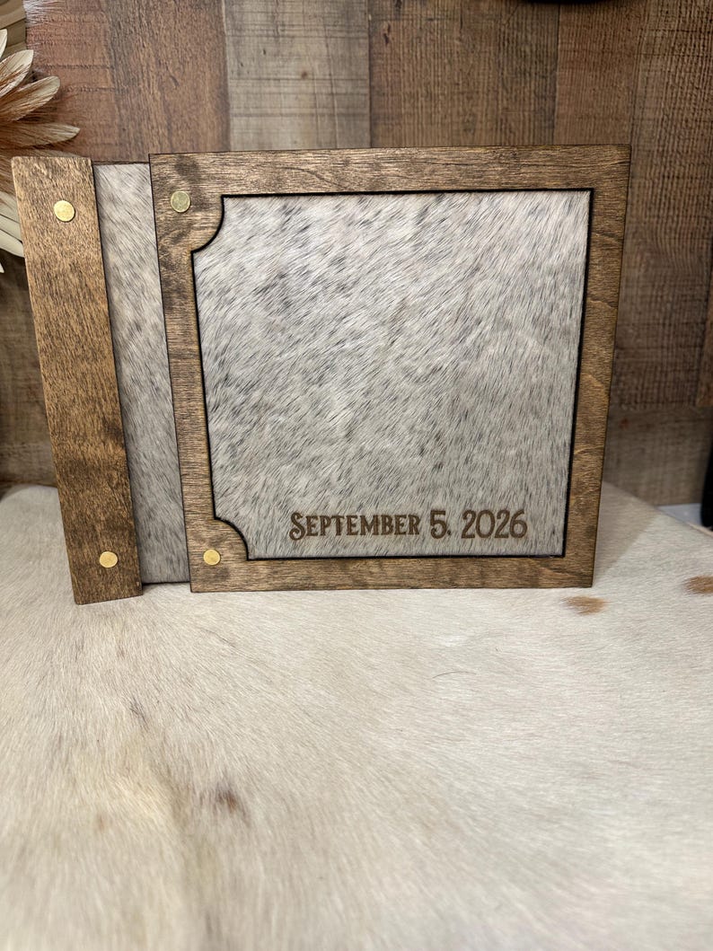 Peut inclure: Un album photo carr&eacute; en bois avec un cadre teint&eacute; brun fonc&eacute; et une incrustation en peau de vache tachet&eacute;e grise et blanche. La couverture pr&eacute;sente le texte grav&eacute; SEPTEMBER 5, 2026, et quatre petits rivets m&eacute;talliques couleur laiton.