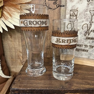 Peut inclure: Deux verres transparents, une chope à bière haute et un verre droit, ornés d'une bande en cuir marron à bords festonnés et d'une pièce centrale en peau de vache blanche et marron portant les inscriptions GROOM et BRIDE.
