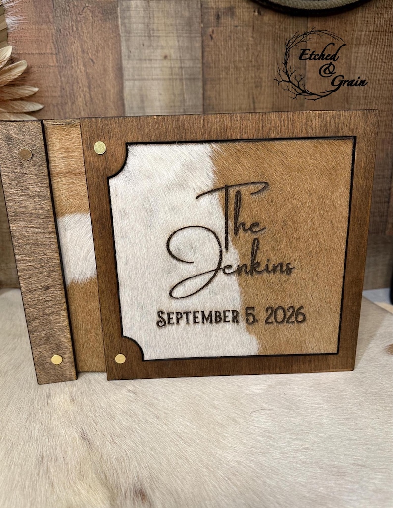 Peut inclure: Une bo&icirc;te carr&eacute;e en bois avec un couvercle coulissant dot&eacute; d'une incrustation en peau de vache marron et blanche. Le couvercle est grav&eacute; avec le texte 'The Jenkins' en &eacute;criture cursive et 'SEPTEMBER 5, 2026' en lettres capitales, avec quatre rivets en m&eacute;tal dor&eacute;.