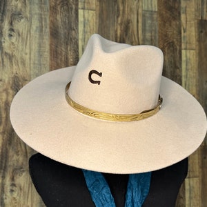 K&ouml;nnte beinhalten: Ein hellbeiger Filz-Fedora-Hut mit einem dunkelbraunen Hufeisen-Emblem, das auf die Vorderseite der Krone gestickt ist. Der Hut ist mit einem metallisch goldfarbenen Band verziert, das ein gepr&auml;gtes Muster und eine kleine seitliche Schnalle aufweist.
