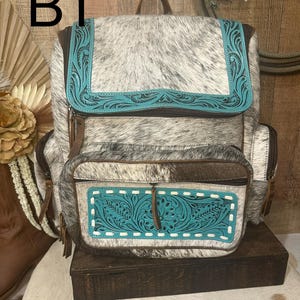 Op de afbeelding: Een rugzak van grijs en wit koeienhuid met turquoise lederen accenten en in reli&euml;f gemaakte bloemmotieven. De tas heeft een bovenflap, een voorvak met rits en witte stiksels, en bruine lederen ritstrekkers. De tekst B1 is zichtbaar.