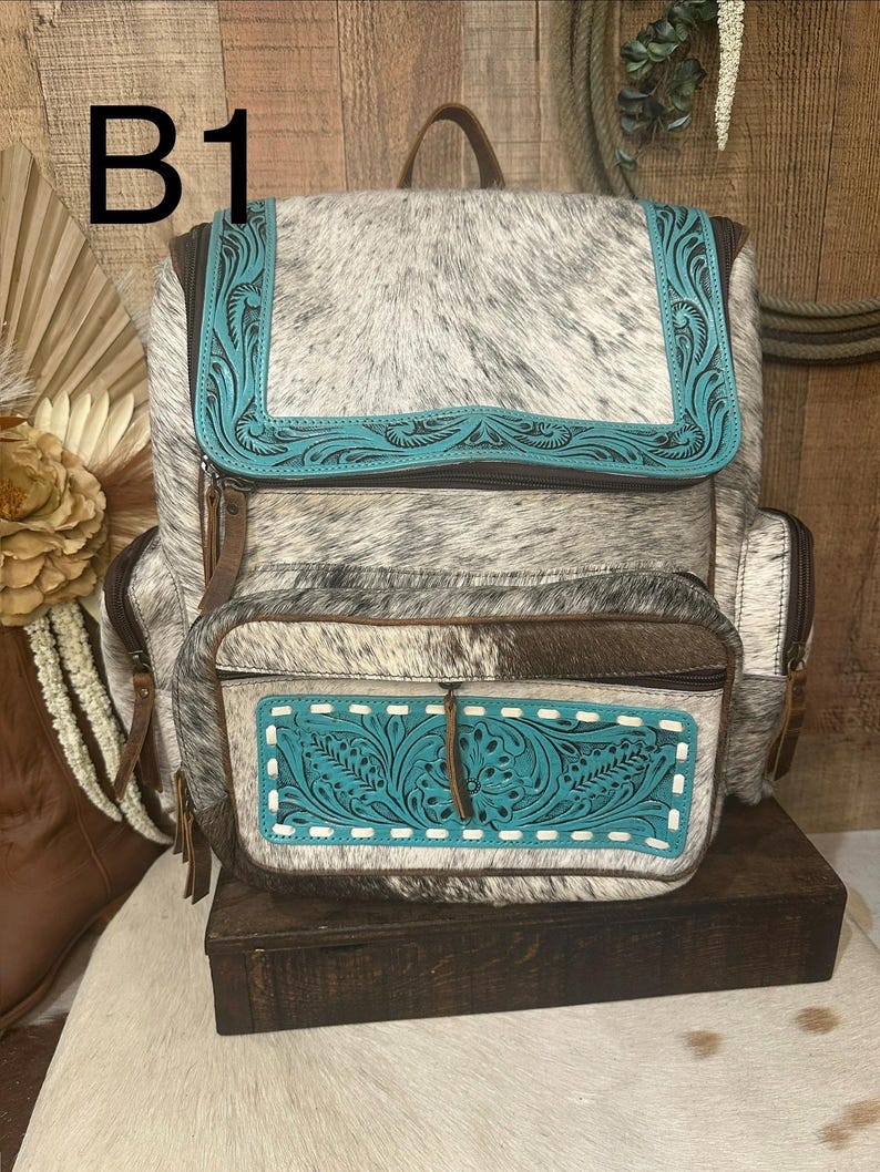 Op de afbeelding: Een rugzak van grijs en wit koeienhuid met turquoise lederen accenten en in reli&euml;f gemaakte bloemmotieven. De tas heeft een bovenflap, een voorvak met rits en witte stiksels, en bruine lederen ritstrekkers. De tekst B1 is zichtbaar.