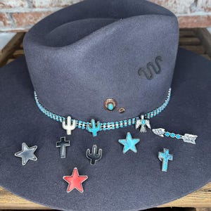 May include: A dark gray felt cowboy hat with a black leather band and turquoise and silver embellishments. The hat has a black felt band with the letters "WC" embroidered on it. The hat is decorated with a variety of charms, including stars, crosses, cacti, and an arrow.