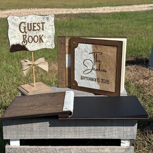 Peut inclure: Un panneau livre d'or avec une texture peau de vache et le texte GUEST BOOK, accompagn&eacute; d'un livre d'or carr&eacute; assorti avec le texte The Jenkins SEPTEMBER 5 2026 sur une couverture en peau de vache, et un livre ouvert aux pages noires.
