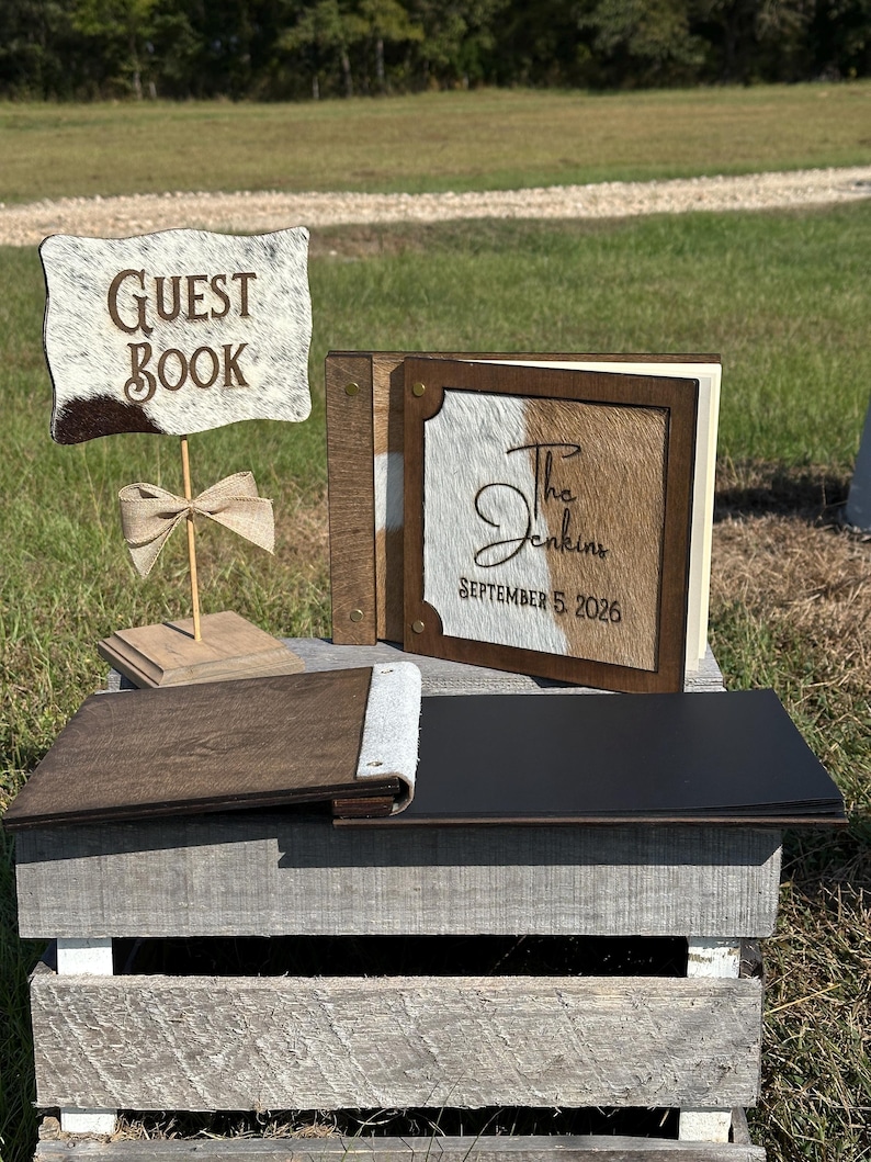 Peut inclure: Un panneau livre d'or avec une texture peau de vache et le texte GUEST BOOK, accompagn&eacute; d'un livre d'or carr&eacute; assorti avec le texte The Jenkins SEPTEMBER 5 2026 sur une couverture en peau de vache, et un livre ouvert aux pages noires.