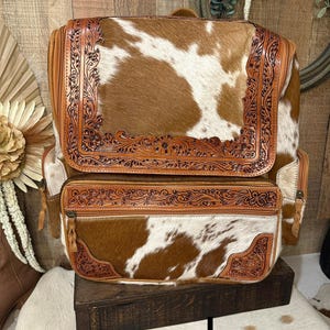 May include: A brown and white cowhide backpack featuring tan leather trim with intricate floral tooling. The bag includes a large top flap and a front zippered pocket, both accented with detailed embossed leather borders and leather zipper pulls.