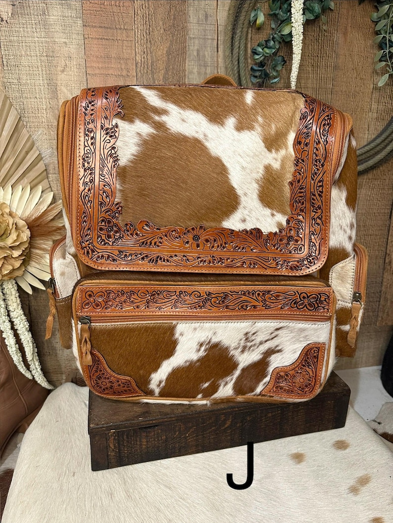 May include: A brown and white cowhide backpack featuring tan leather trim with intricate floral tooling. The bag includes a large top flap and a front zippered pocket, both accented with detailed embossed leather borders and leather zipper pulls.