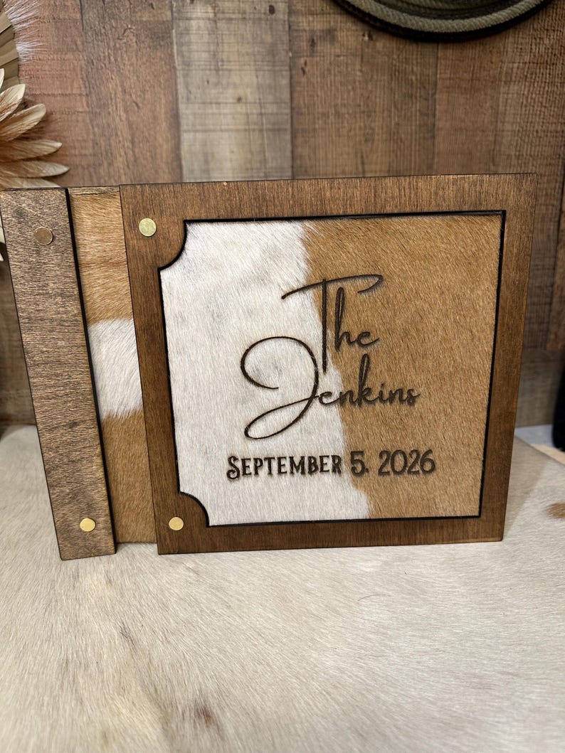 Peut inclure: Un album photo rectangulaire en bois avec une incrustation en peau de vache aux taches brunes et blanches. La couverture est grav&eacute;e avec le texte The Jenkins et la date SEPTEMBER 5, 2026. L'album est fix&eacute; par de petits rivets en m&eacute;tal dor&eacute;.