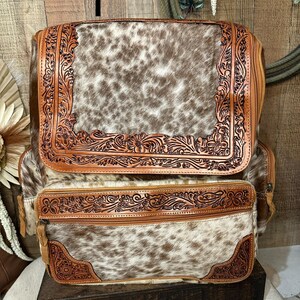 May include: A backpack featuring a brown and white cowhide pattern with tan leather trim. The leather sections are decorated with intricate floral tooling. The bag includes a large top flap and a smaller front pocket, both secured with zippers.