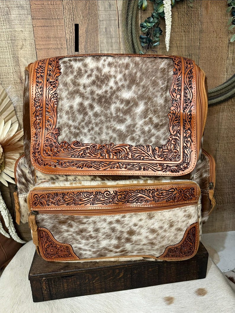 May include: A backpack featuring a brown and white cowhide pattern with tan leather trim. The leather sections are decorated with intricate floral tooling. The bag includes a large top flap and a smaller front pocket, both secured with zippers.