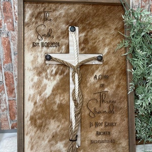 May include: A wooden framed sign with a brown and white cowhide background. A white rope is woven into a cross shape with the text "The Smiths Est. 11.02.2024" and "A Cord of Three Strands Is Not Easily Broken Ecclesiastes 4:12".