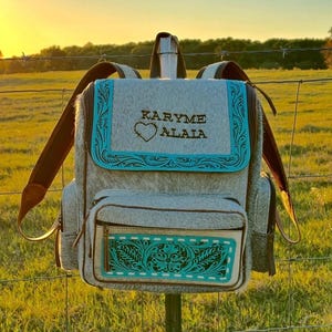 May include: A gray and white cowhide backpack featuring a top flap with the text KARYME ALALA and a heart icon. The bag includes turquoise leather trim with embossed floral patterns, brown leather straps, and a front pocket with matching trim.