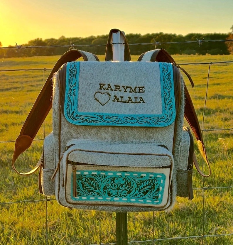 May include: A gray and white cowhide backpack featuring a top flap with the text KARYME ALALA and a heart icon. The bag includes turquoise leather trim with embossed floral patterns, brown leather straps, and a front pocket with matching trim.