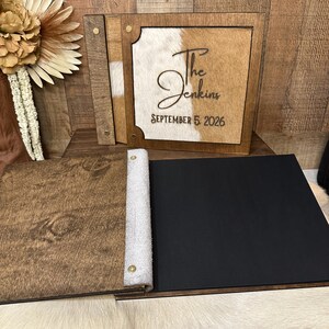 Peut inclure: Un album photo en bois avec une couverture en peau de vache portant le texte The Jenkins et SEPTEMBER 5, 2026. L'album pr&eacute;sente un cadre en bois fonc&eacute;, une incrustation en peau blanche et brune, et une section ouverte avec des pages noires.