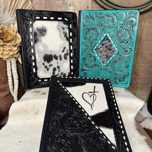 May include: Three rectangular leather binders featuring intricate floral tooling. One binder is black with a white and black cowhide center panel. Another is turquoise with a diamond-shaped cowhide inlay. The third is black with a diagonal cowhide panel displaying a black heart and cross symbol.