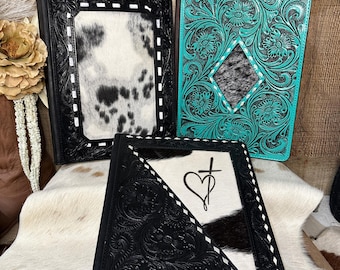 Puede incluir: Tres carpetas rectangulares de cuero con intrincados grabados florales. Una es negra con un panel central de piel de vaca blanca y negra. Otra es turquesa con una incrustación de piel de vaca en forma de diamante. La tercera es negra con un panel diagonal de piel de vaca que muestra un símbolo de corazón y cruz.