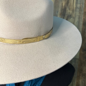K&ouml;nnte beinhalten: Ein hellbeiger Fedora-Hut mit breiter Krempe und strukturierter Krone. Der Hut ist mit einem metallisch goldfarbenen Band akzentuiert, das um die Basis verl&auml;uft und ein komplexes eingraviertes Muster sowie ein glattes, ovales Metalldetail zeigt.