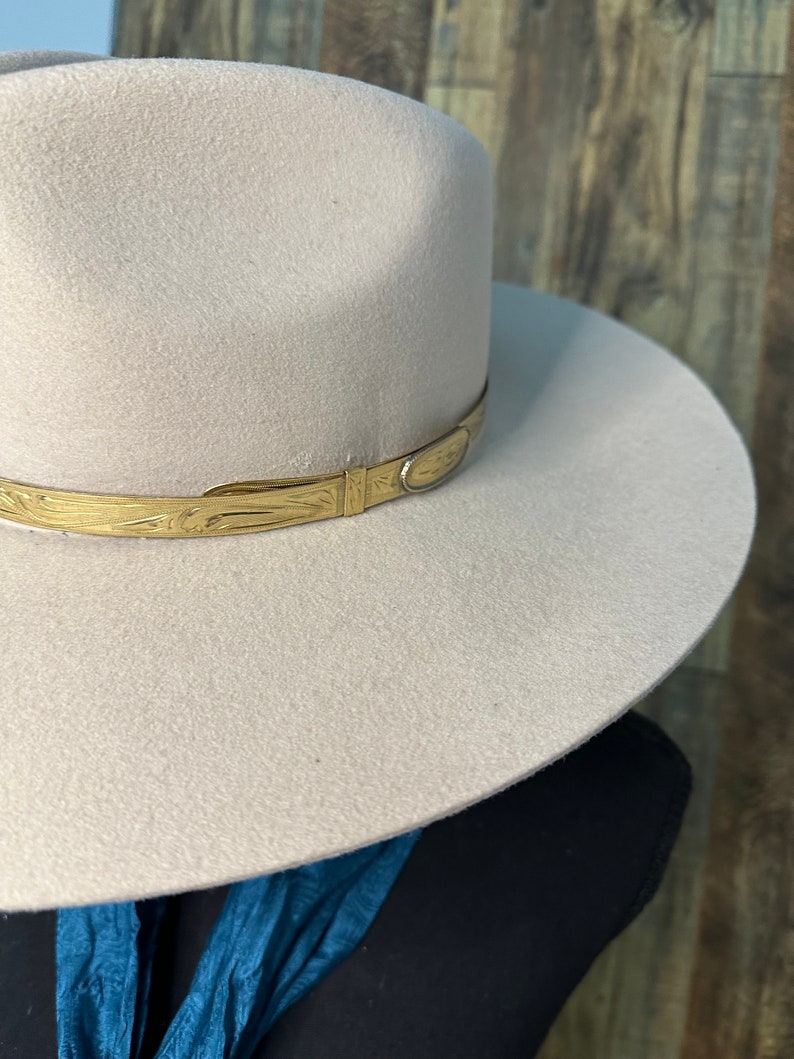 K&ouml;nnte beinhalten: Ein hellbeiger Fedora-Hut mit breiter Krempe und strukturierter Krone. Der Hut ist mit einem metallisch goldfarbenen Band akzentuiert, das um die Basis verl&auml;uft und ein komplexes eingraviertes Muster sowie ein glattes, ovales Metalldetail zeigt.