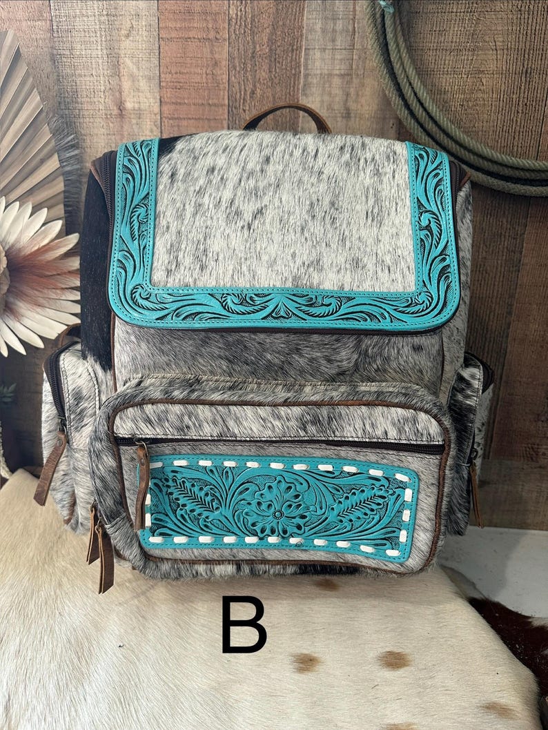 May include: A backpack crafted from gray and white speckled cowhide leather, featuring a top flap and front pocket accented with bright turquoise leather panels. The turquoise sections are embossed with intricate floral scrollwork and white stitching.