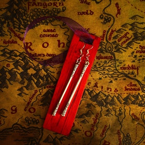 May include: A pair of silver dangle earrings with a textured design, displayed on a red background. The earrings are shown against a map of Middle-earth, a fictional world from the Lord of the Rings.