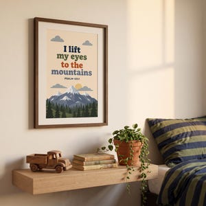 May include: A framed wall art print featuring a mountain landscape illustration with a forest, clouds, and a sun. The text reads: I lift my eyes to the mountains PSALM 121:1. The design uses a palette of blue, green, orange, and cream colours.