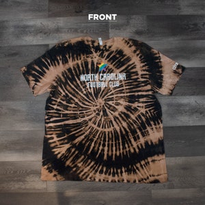 以下が含まれることがあります： 白の星と「NORTH CAROLINA FOOTBALL CLUB」の文字が入った黒と茶色のタイダイ染めTシャツです。