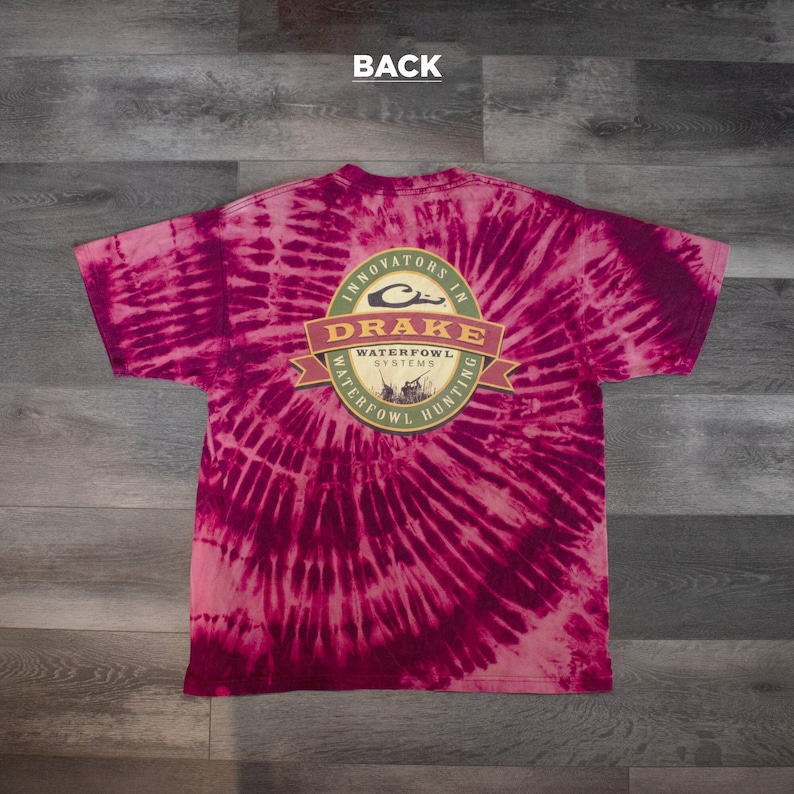 May include: A pink and white tie-dye t-shirt with a circular logo featuring the text "Drake Waterfowl Systems" and "Innovators in Waterfowl Hunting".