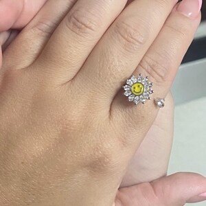 May include: A silver ring with a white daisy design featuring a yellow smiley face in the center. The ring is on a person's finger.