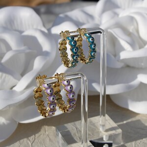 May include: Two pairs of gold hoop earrings with a heart-shaped design. One pair has turquoise blue evil eyes and the other pair has lavender evil eyes.