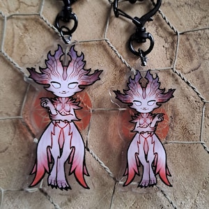 Peut inclure: Deux porte-clés en acrylique transparent représentant une créature rose et blanche avec un contour noir. La créature a une grosse tête avec une couronne pointue et de longs cheveux flottants. Les porte-clés sont attachés à des fermoirs en forme de cœur noirs.