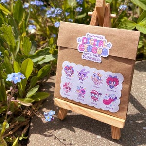 Pode incluir: Um envelope de papel castanho num pequeno cavalete de madeira. O envelope apresenta um autocolante com a inscri&ccedil;&atilde;o Fanmade Digital Circus Keychain Blind Bag e um segundo autocolante com as personagens Pomni, Jax, Kinger, Ragatha, Gangle, Zooble, Caine e Bubble.