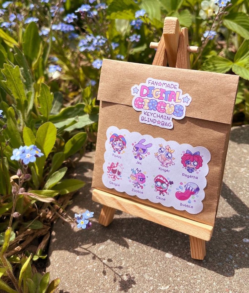 Pode incluir: Um envelope de papel castanho num pequeno cavalete de madeira. O envelope apresenta um autocolante com a inscri&ccedil;&atilde;o Fanmade Digital Circus Keychain Blind Bag e um segundo autocolante com as personagens Pomni, Jax, Kinger, Ragatha, Gangle, Zooble, Caine e Bubble.